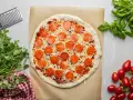 Surowa pizza z sosem, serem i okrągłymi plasterkami peperoni, gotowa do pieczenia. Obok świeża bazylia i pomidorki.