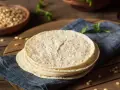 Stos świeżych, okrągłych placków, czyli co to jest tortilla, leży na niebieskiej serwetce. W tle widać miskę z kukurydzą.
