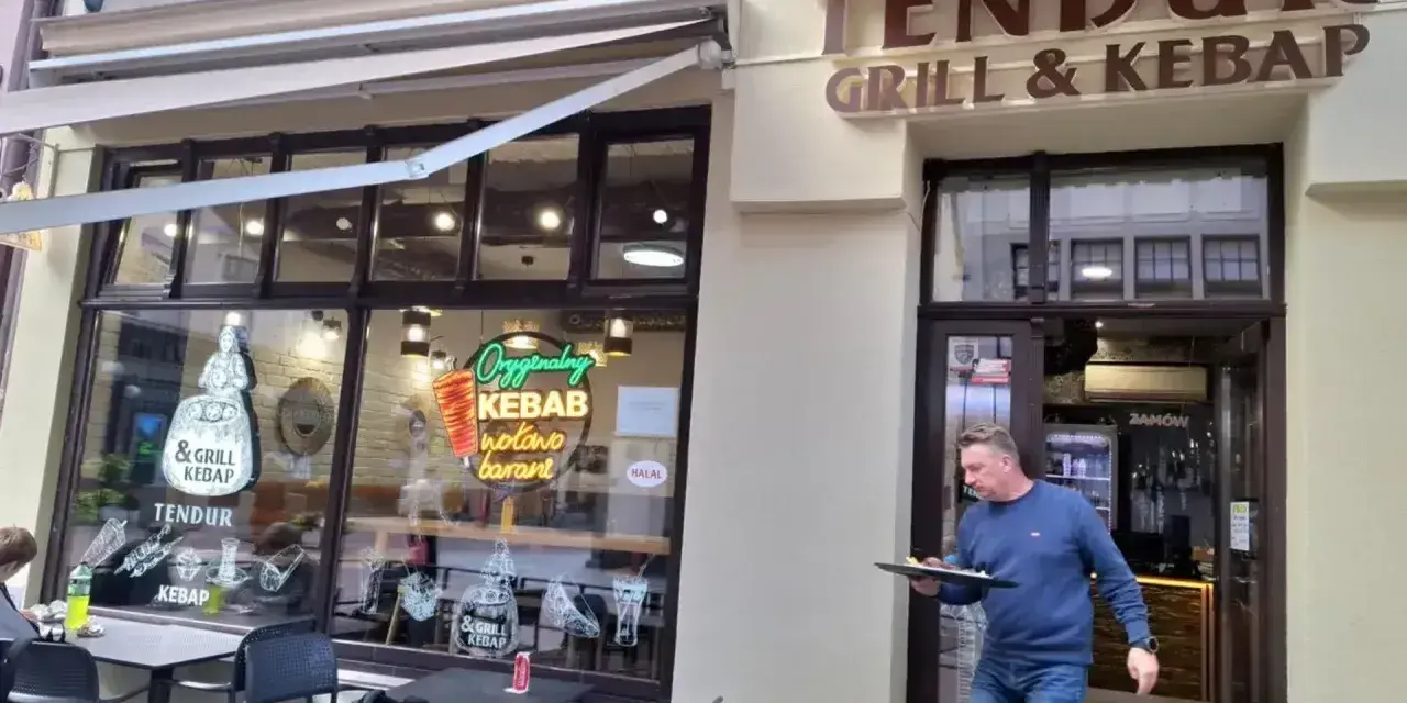 Mężczyzna wychodzi z lokalu "Tendur Grill & Kebap", niosąc talerz. W witrynie neonowy napis "Oryginalny Kebab".