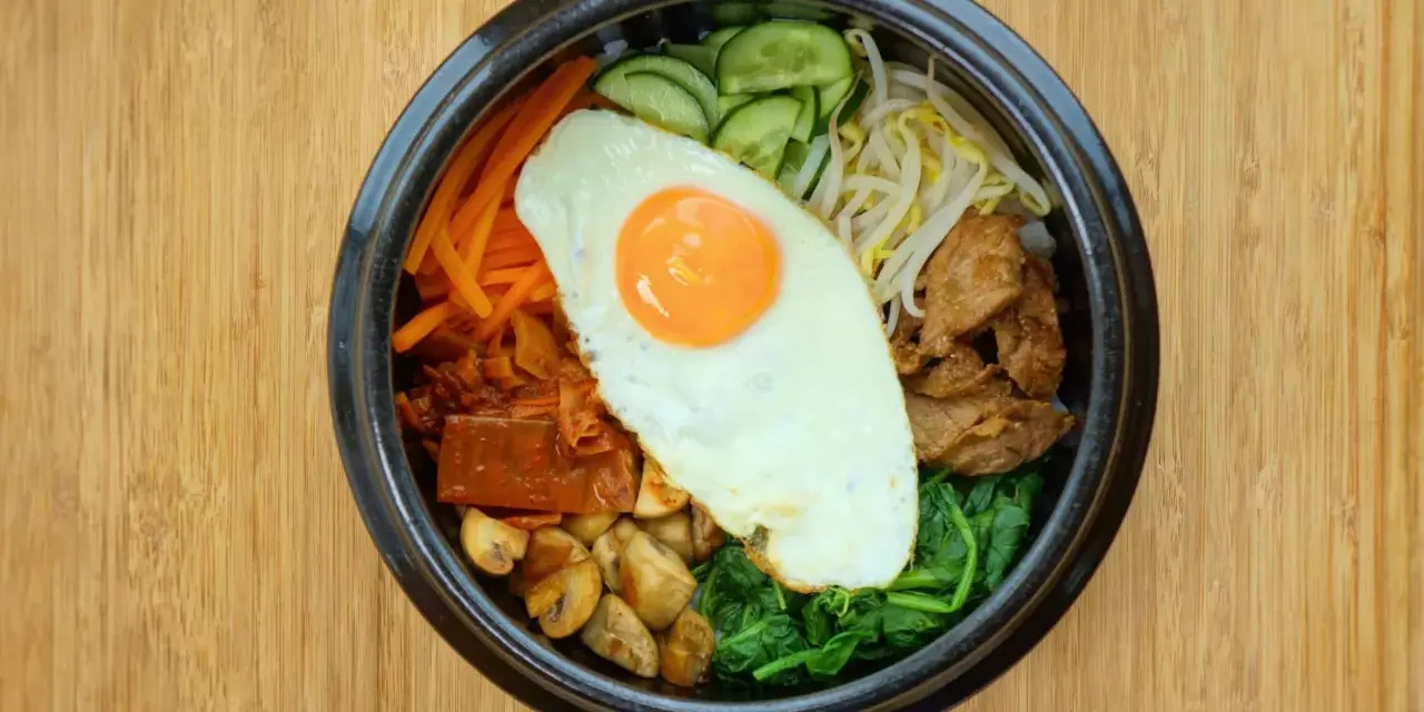 Kolorowy bibimbap z jajkiem sadzonym, wołowiną, warzywami i grzybami na drewnianym tle.