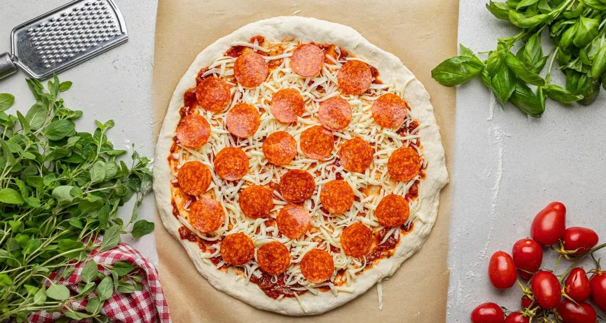 Surowa pizza z sosem, serem i okrągłymi plasterkami peperoni, gotowa do pieczenia. Obok świeża bazylia i pomidorki.