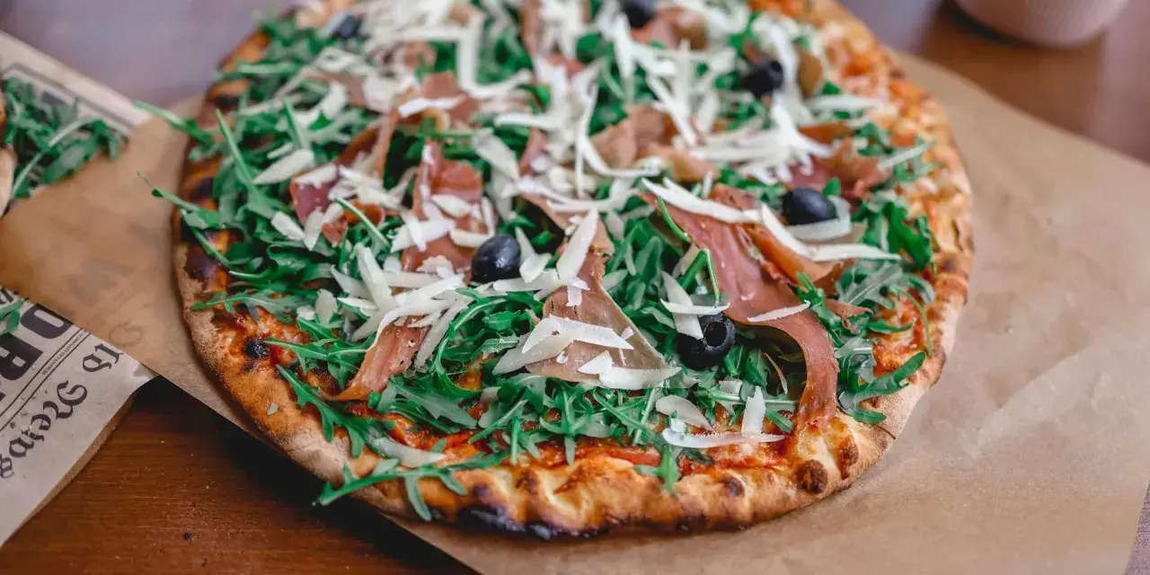 Pyszna pizza z rukolą, oliwkami, parmezanem i cienkimi plastrami prosciutto.