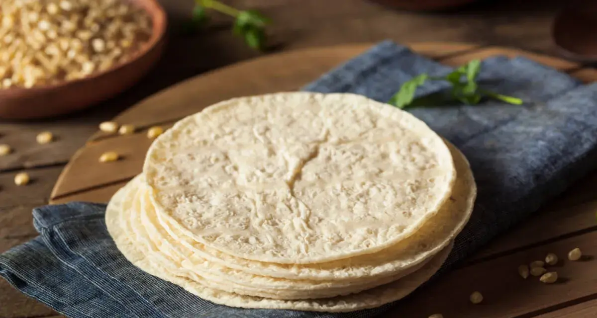 Stos świeżych, okrągłych placków, czyli co to jest tortilla, leży na niebieskiej serwetce. W tle widać miskę z kukurydzą.