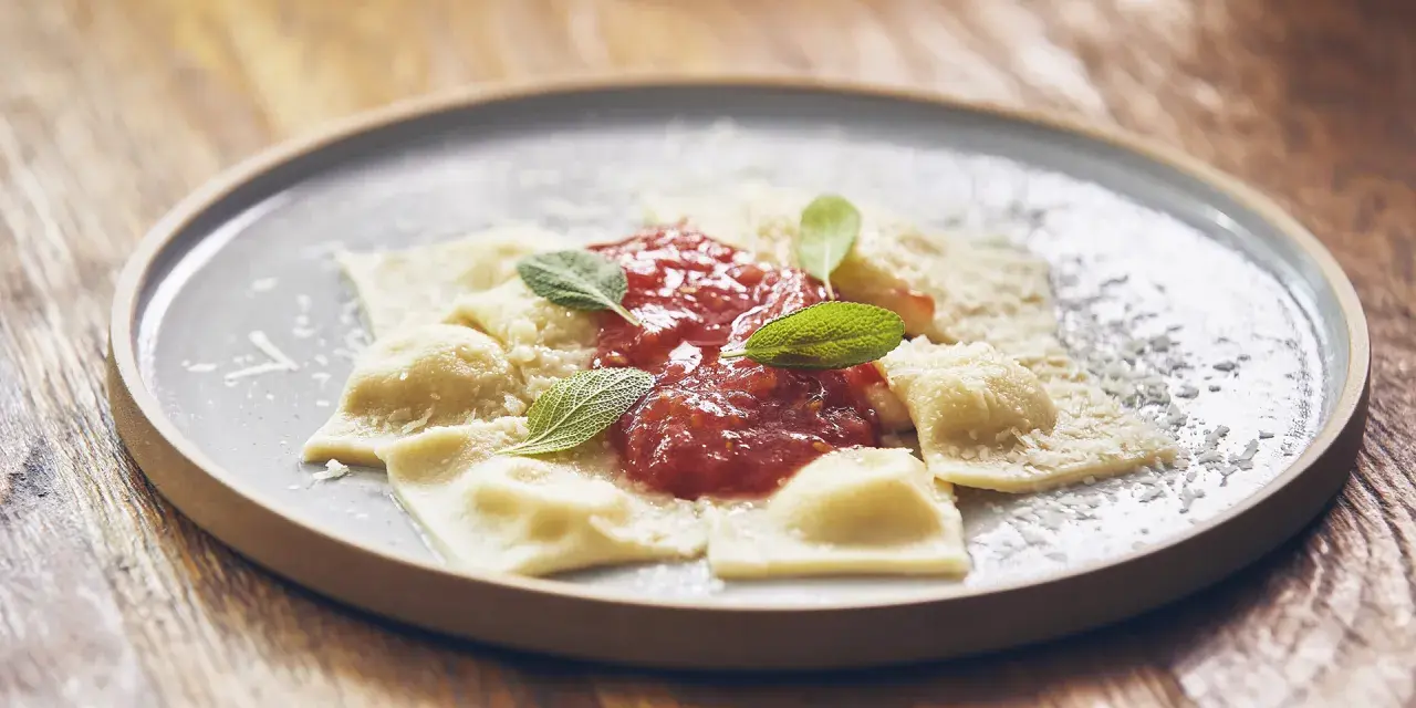 Pyszne raviolini z sosem pomidorowym i parmezanem, ozdobione listkami szałwii, na szarym talerzu.