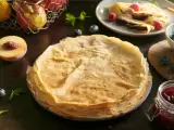 Stos cienkich, złocistych naleśników po francusku, obok brzoskwini, jagód i słoiczka dżemu.