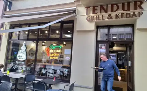 Mężczyzna wychodzi z lokalu "Tendur Grill & Kebap", niosąc talerz. W witrynie neonowy napis "Oryginalny Kebab".