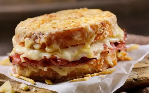 Pyszny croque madame z rozpływającym się serem i szynką, podany na papierze na drewnianej desce.