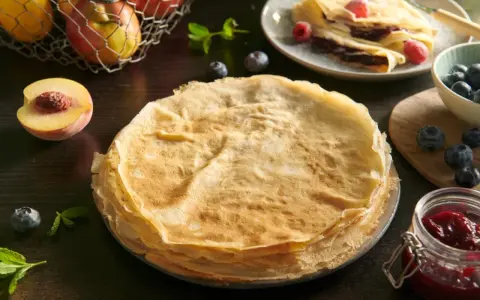 Stos cienkich, złocistych naleśników po francusku, obok brzoskwini, jagód i słoiczka dżemu.