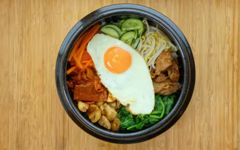Kolorowy bibimbap z jajkiem sadzonym, wołowiną, warzywami i grzybami na drewnianym tle.