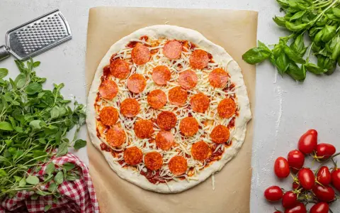 Surowa pizza z sosem, serem i okrągłymi plasterkami peperoni, gotowa do pieczenia. Obok świeża bazylia i pomidorki.