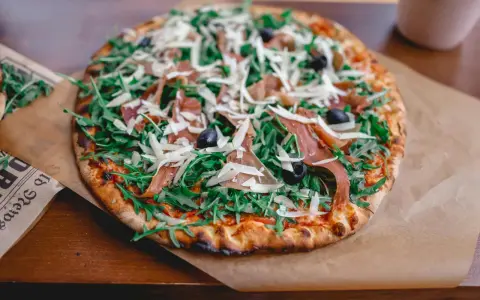 Pyszna pizza z rukolą, oliwkami, parmezanem i cienkimi plastrami prosciutto.