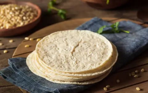 Stos świeżych, okrągłych placków, czyli co to jest tortilla, leży na niebieskiej serwetce. W tle widać miskę z kukurydzą.