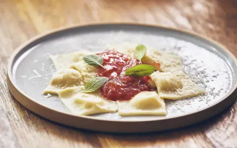 Pyszne raviolini z sosem pomidorowym i parmezanem, ozdobione listkami szałwii, na szarym talerzu.