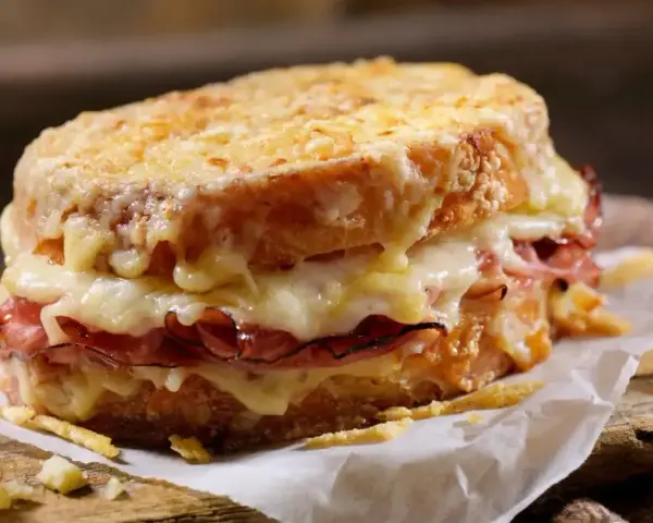 Pyszny croque madame z rozpływającym się serem i szynką, podany na papierze na drewnianej desce.