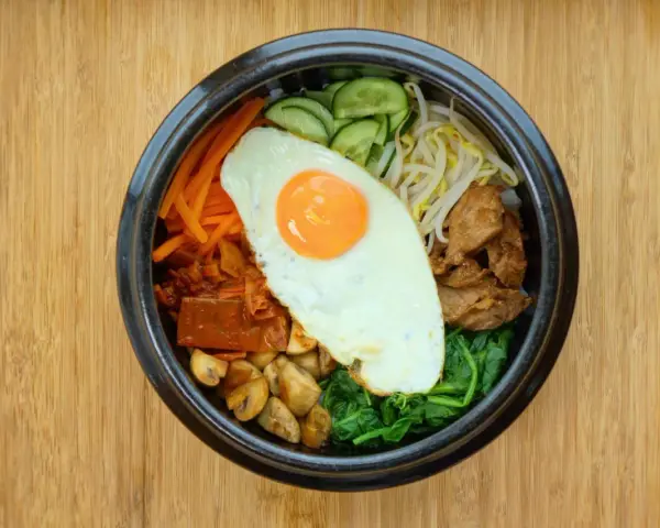Kolorowy bibimbap z jajkiem sadzonym, wołowiną, warzywami i grzybami na drewnianym tle.