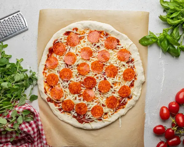 Surowa pizza z sosem, serem i okrągłymi plasterkami peperoni, gotowa do pieczenia. Obok świeża bazylia i pomidorki.