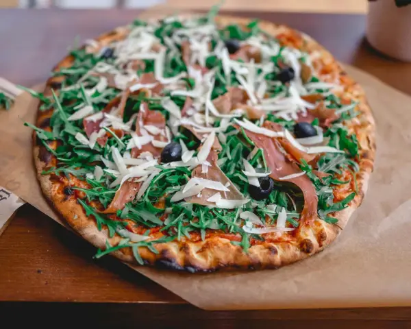 Pyszna pizza z rukolą, oliwkami, parmezanem i cienkimi plastrami prosciutto.