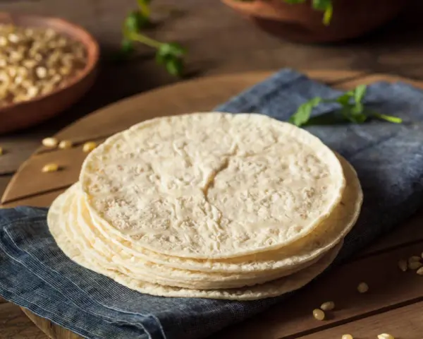 Stos świeżych, okrągłych placków, czyli co to jest tortilla, leży na niebieskiej serwetce. W tle widać miskę z kukurydzą.