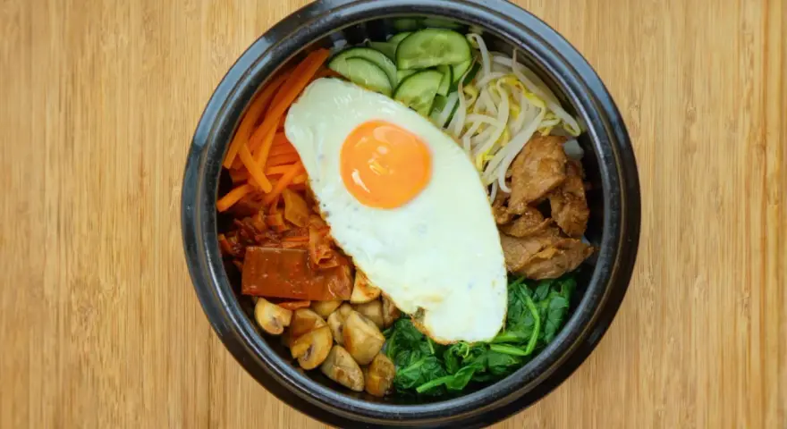 Kolorowy bibimbap z jajkiem sadzonym, wołowiną, warzywami i grzybami na drewnianym tle.
