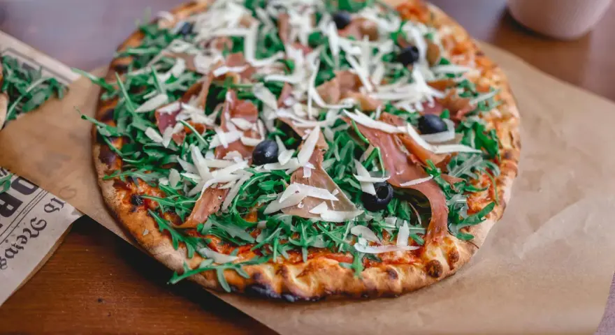 Pyszna pizza z rukolą, oliwkami, parmezanem i cienkimi plastrami prosciutto.