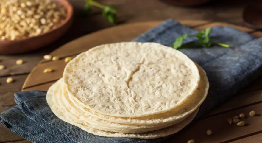 Stos świeżych, okrągłych placków, czyli co to jest tortilla, leży na niebieskiej serwetce. W tle widać miskę z kukurydzą.