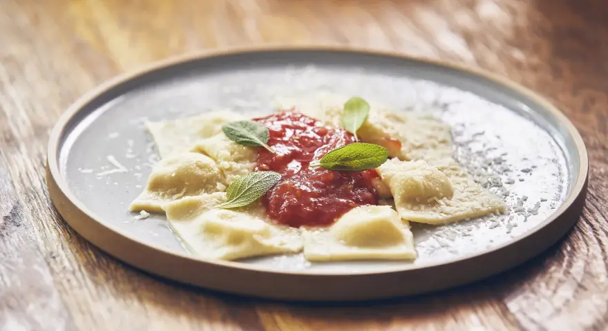 Pyszne raviolini z sosem pomidorowym i parmezanem, ozdobione listkami szałwii, na szarym talerzu.