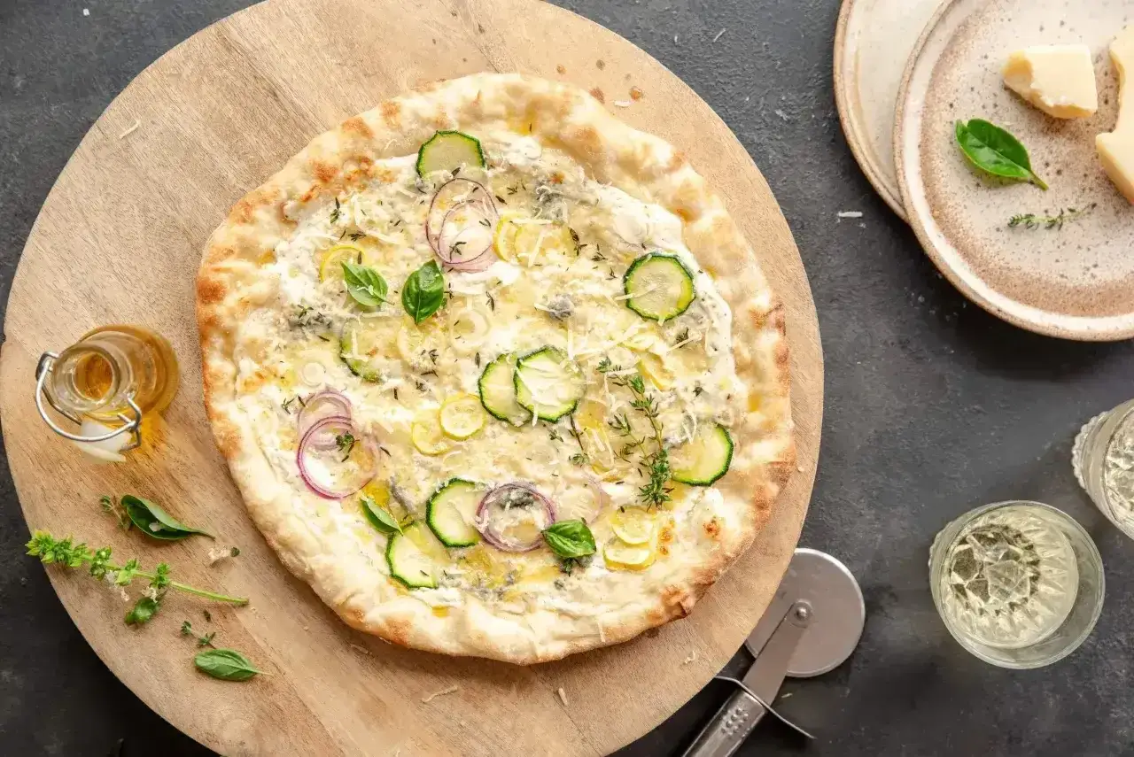 Apetyczna pizza bianca z cukinią, cebulą i ziołami, podana na drewnianej desce. Obok buteleczka oliwy i kieliszki z winem.