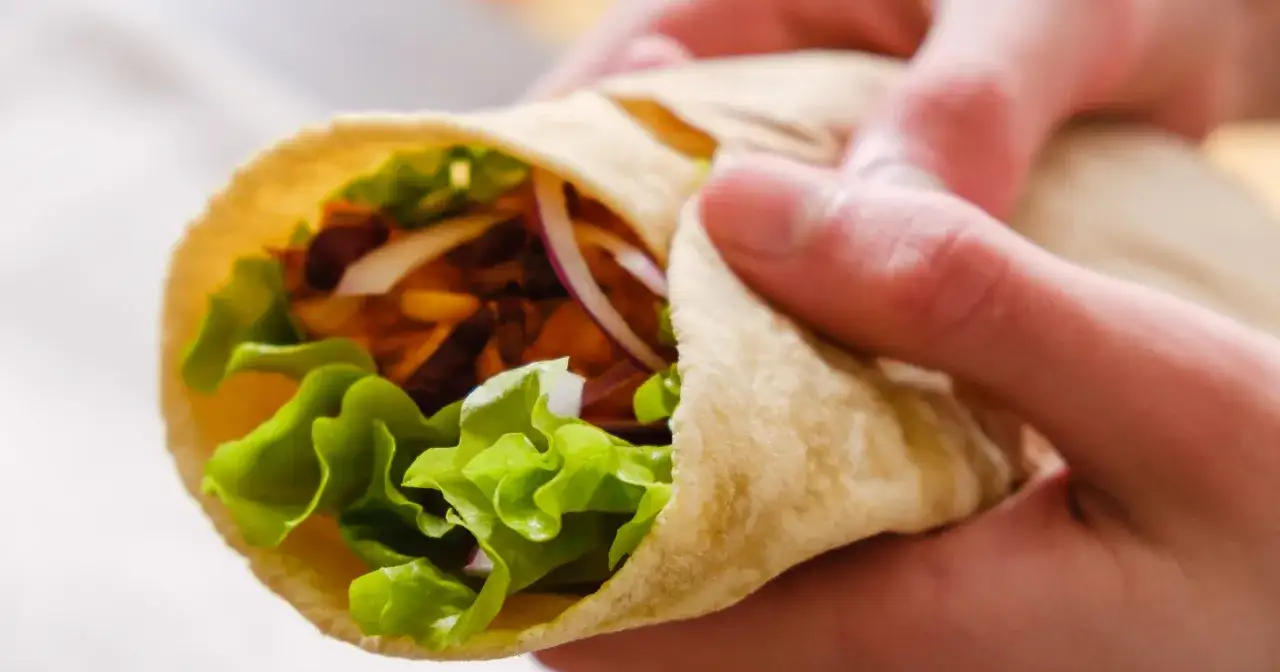 Ręce trzymają zawiniętą tortillę z sałatą, cebulą i mięsem. To pyszne danie, które pokazuje, co to jest tortilla.