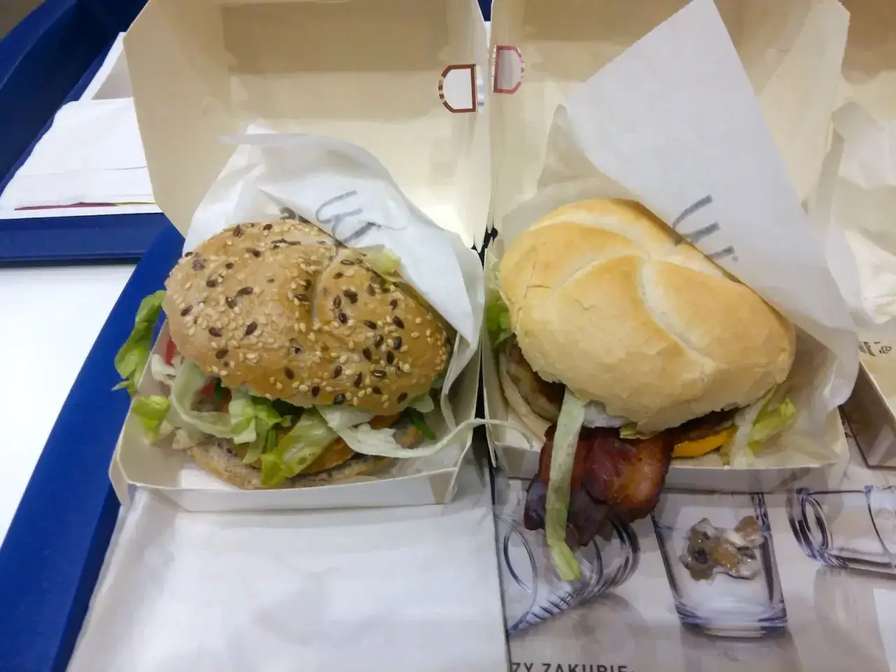 Dwa burgery w papierowych opakowaniach. Jeden z bułką z sezamem, drugi z jasną bułką i bekonem. Pyszna kajzerka wieprzowa deluxe, ile kcal?