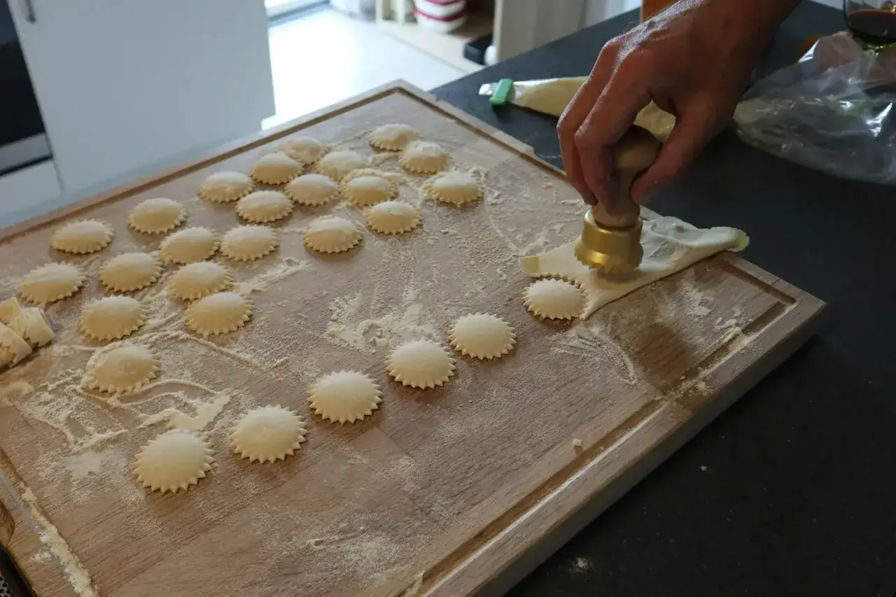 Ręcznie robione ravioli co to? Na drewnianej desce powstają okrągłe pierożki z ciasta, posypane mąką. Dłoń dociska złote wykrawaczki.