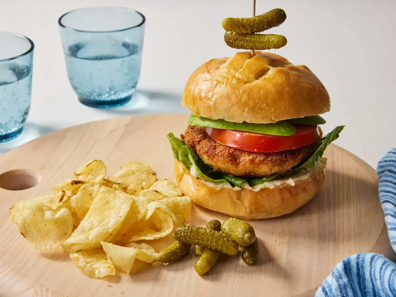 Pyszny chicken burger z chrupiącym kotletem, świeżymi warzywami i ogórkami. Obok frytki i dwa szklanki wody. Idealny posiłek, który nie przekroczy Twoich założeń kalorycznych.