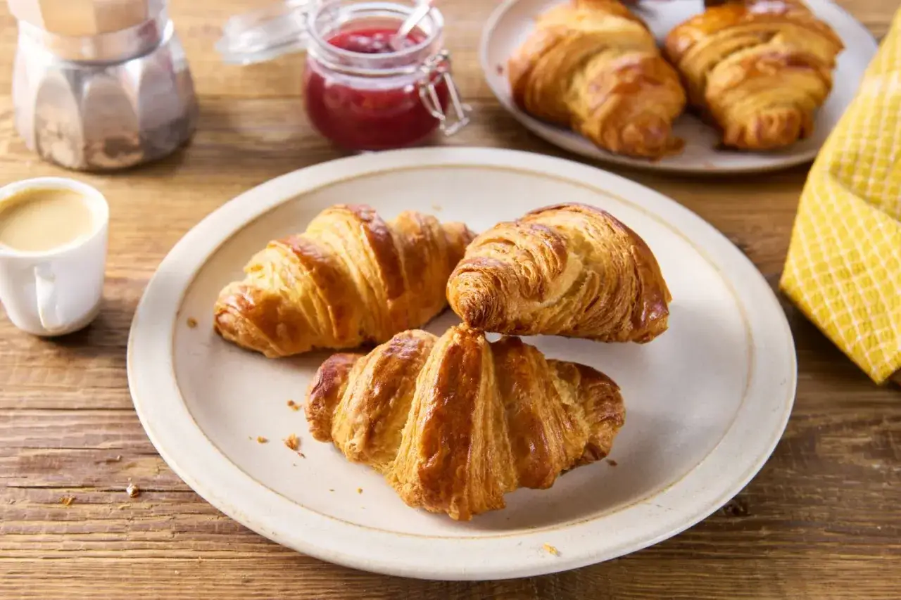 Złociste, chrupiące croissanty na talerzu, obok filiżanka kawy i słoiczek dżemu. Idealne śniadanie.