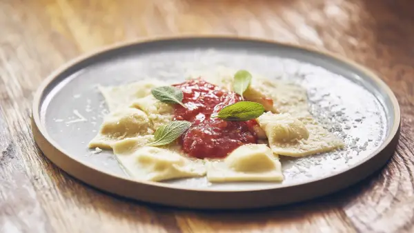 Pyszne raviolini z sosem pomidorowym i parmezanem, ozdobione listkami szałwii, na szarym talerzu.